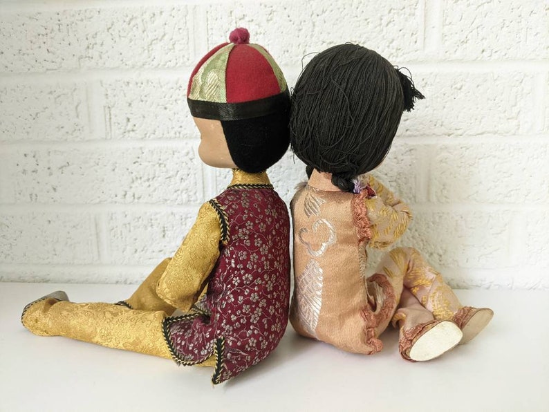 Op de afbeelding: Twee vintage poppen, rug aan rug, gekleed in traditionele Aziatische kleding. De pop links draagt een rood-gouden hoed en een bordeauxrode vest over een gouden outfit. De pop rechts heeft zwart haar en een perzikkleurige outfit met gouden accenten.