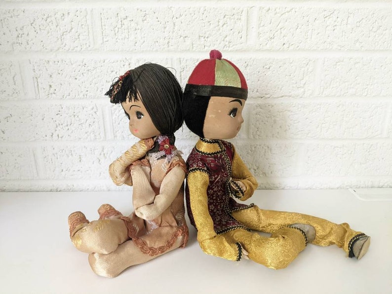 Op de afbeelding: Twee vintage Aziatische poppen, een meisje en een jongen, zitten rug aan rug. De meisjespop draagt een perzikkleurige jurk met bloemaccenten. De jongenspop is gekleed in een goud-rode outfit met een bijpassende hoed. Beide poppen hebben zwart haar en geschilderde gezichtskenmerken.