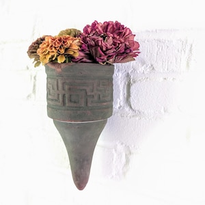 May include: A wall-mounted, dark gray ceramic vase with a geometric pattern, holding a bouquet of dried flowers in shades of orange, yellow, and burgundy. The vase has a cone-shaped base and is mounted on a white brick wall.