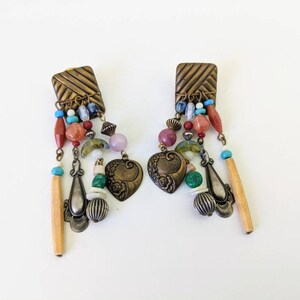 May include: A pair of dangle earrings with a square-shaped top and a variety of colourful beads, including red, green, blue, and purple. The earrings feature a heart-shaped charm and a long, thin wooden bead.