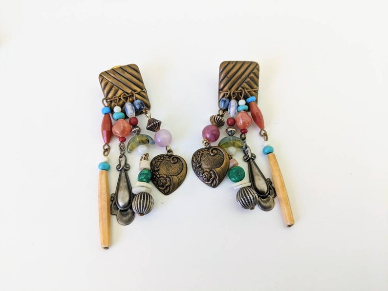 May include: A pair of dangle earrings with a square-shaped top and a variety of colourful beads, including red, green, blue, and purple. The earrings feature a heart-shaped charm and a long, thin wooden bead.