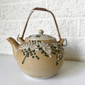 May include: A brown ceramic teapot with a floral design in white and green. The teapot has a woven brown handle and a lid with a small knob.