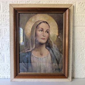 May include: A framed portrait of a woman with a golden halo, wearing a blue shawl and a white dress. The artwork is in a wooden frame with a textured, off-white border. The painting has a vintage aesthetic.