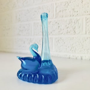May include: A blue glass swan vase with a tall neck. The swan is sitting on a base with a textured surface.