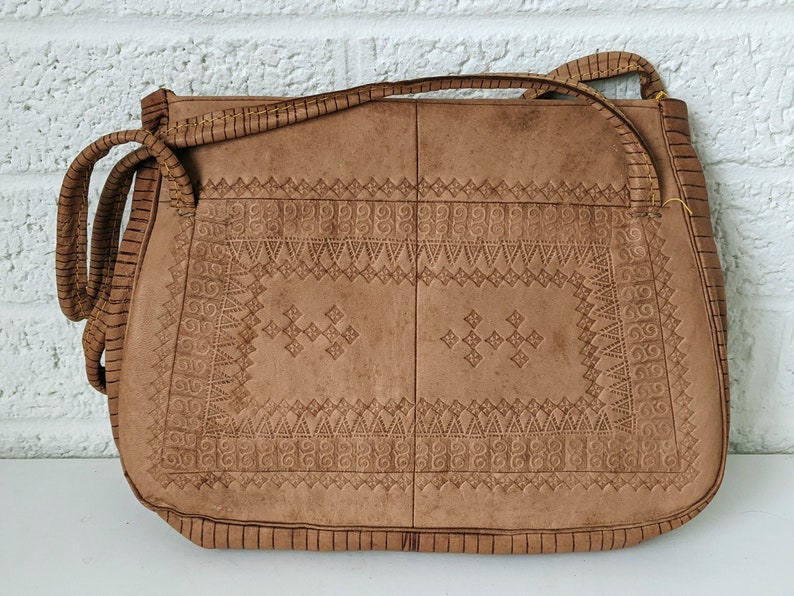 Puede incluir: Un bolso de hombro de cuero marr&oacute;n con intrincados patrones en relieve. El bolso tiene un cierre de solapa y una correa larga.