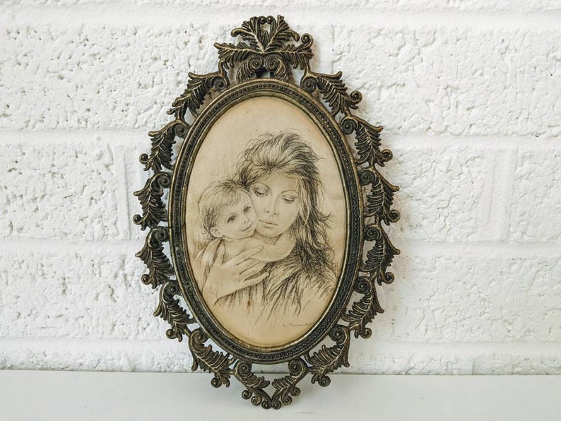 May include: A vintage oval-shaped picture frame with an ornate gold metal design. The frame holds a black and white drawing of a mother and child.