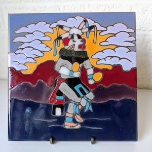 Puede incluir: Un azulejo de cerámica colorido que representa a un bailarín nativo americano con un traje tradicional. El bailarín está rodeado de nubes y una puesta de sol. El azulejo es una mezcla vibrante de azul, rojo, amarillo y blanco.
