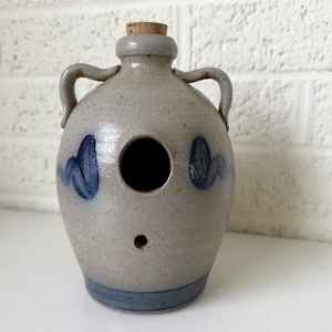 May include: A gray stoneware pottery birdhouse with blue and white glaze. The birdhouse has a round opening and a cork stopper on top.