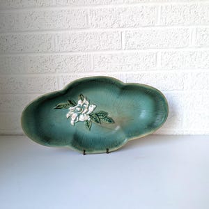Puede incluir: Un plato de servir de cer&aacute;mica verde azulado con un dise&ntilde;o de flor blanca en relieve. El plato tiene un borde festoneado y tiene forma de hoja.