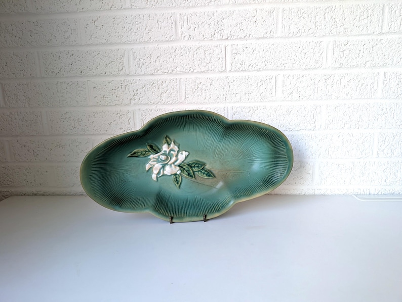 Puede incluir: Un plato de servir de cer&aacute;mica verde azulado con un dise&ntilde;o de flor blanca en relieve. El plato tiene un borde festoneado y tiene forma de hoja.