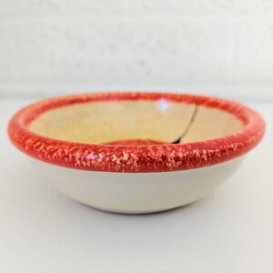 May include: A small, white ceramic bowl with a red rim. The rim has a speckled pattern. The bowl is slightly chipped.