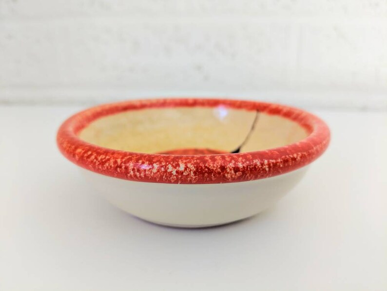 May include: A small, white ceramic bowl with a red rim. The rim has a speckled pattern. The bowl is slightly chipped.