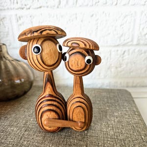 Pode incluir: Um par de estatuetas de madeira com olhos grandes e traços simples. As estatuetas são feitas de madeira em camadas e são pintadas em tons de marrom. A estatueta maior está usando um chapéu.