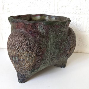 May include: A rustic, green and brown ceramic planter with a wavy rim and three legs. The planter has a textured surface.