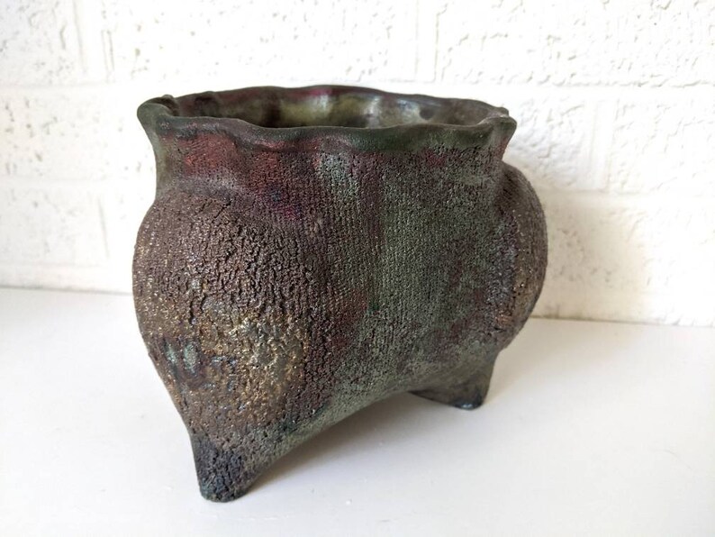 May include: A rustic, green and brown ceramic planter with a wavy rim and three legs. The planter has a textured surface.