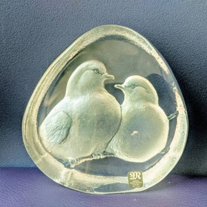 May include: A clear glass paperweight featuring a pair of doves in a sculpted design. The paperweight is oval shaped and has a raised edge. The doves are facing each other and appear to be perched on a branch. The paperweight is signed "Mats Jonasson Sweden".
