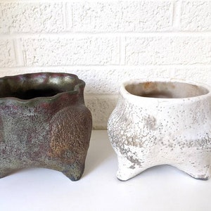 May include: Two ceramic planters with a distressed white and a green and brown glaze. Both planters have three legs and a wide, rounded opening.