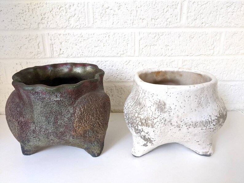 May include: Two ceramic planters with a distressed white and a green and brown glaze. Both planters have three legs and a wide, rounded opening.