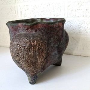 May include: A small, dark brown ceramic planter with a textured surface and three legs. The planter has a slightly flared rim and a deep, rounded body.