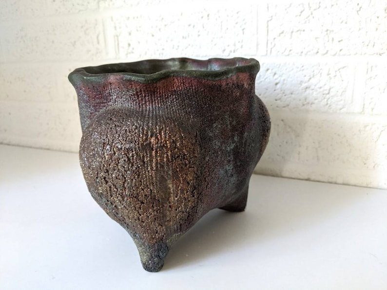 May include: A small, dark brown ceramic planter with a textured surface and three legs. The planter has a slightly flared rim and a deep, rounded body.