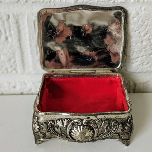 May include: A silver trinket box with an ornate design and a red velvet interior. The box is open and the lid is propped up.