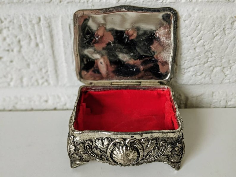 May include: A silver trinket box with an ornate design and a red velvet interior. The box is open and the lid is propped up.
