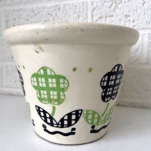 May include: A white ceramic flower pot with a green and black checkered pattern. The pot has a simple design with three flowers and leaves.