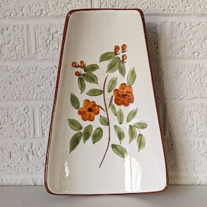 Puede incluir: Un plato de servir de cerámica blanca con un borde marrón. El plato está decorado con un diseño floral pintado a mano con flores naranjas y hojas verdes.