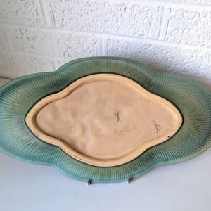 Puede incluir: Un plato de servir de cer&aacute;mica verde con un borde festoneado y un centro elevado. El plato est&aacute; firmado "X. Peart" en la parte inferior.