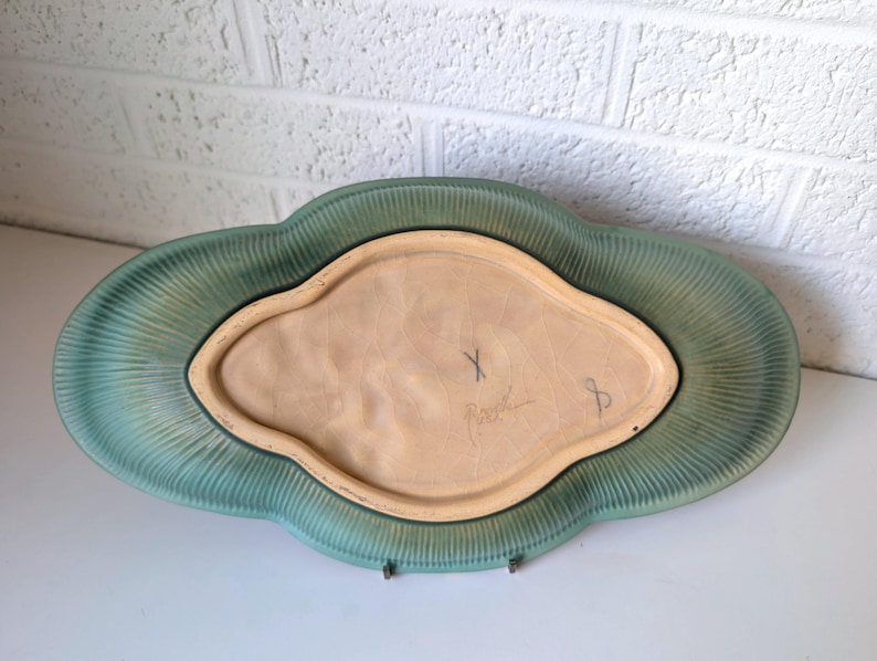 Puede incluir: Un plato de servir de cer&aacute;mica verde con un borde festoneado y un centro elevado. El plato est&aacute; firmado "X. Peart" en la parte inferior.