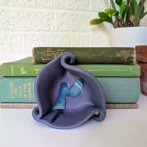 May include: A handcrafted, lavender-colored ceramic dish with a blue accent. The dish has a unique, organic shape and sits in front of a stack of vintage books. The books are various shades of green and have aged covers.