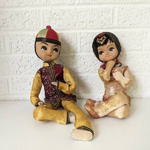 Op de afbeelding: Twee vintage Aziatische poppen. De ene pop is gekleed in een goudkleurige en bordeauxrode outfit met een bijpassende hoed. De andere pop draagt een cr&egrave;mekleurige outfit met gouden accenten. Beide poppen hebben geschilderde gezichten en zitten.