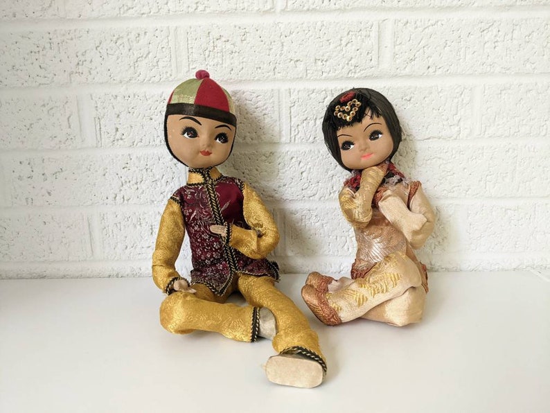 Op de afbeelding: Twee vintage Aziatische poppen. De ene pop is gekleed in een goudkleurige en bordeauxrode outfit met een bijpassende hoed. De andere pop draagt een cr&egrave;mekleurige outfit met gouden accenten. Beide poppen hebben geschilderde gezichten en zitten.