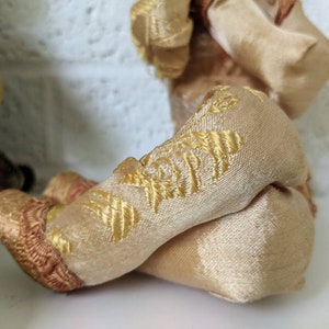 Op de afbeelding: Close-up van de benen van een vintage pop. De benen zijn gemaakt van een zachte, beige stof met gouden borduursels. De voeten zijn een donkerdere bruintint. De benen zijn gekruist.