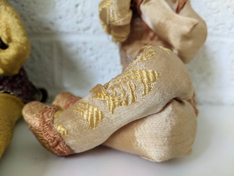 Op de afbeelding: Close-up van de benen van een vintage pop. De benen zijn gemaakt van een zachte, beige stof met gouden borduursels. De voeten zijn een donkerdere bruintint. De benen zijn gekruist.