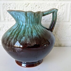 May include: A brown and green glazed ceramic pitcher with a flared lip and a handle. The pitcher has a mottled glaze with shades of brown, green, and blue.