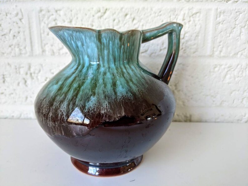 May include: A brown and green glazed ceramic pitcher with a flared lip and a handle. The pitcher has a mottled glaze with shades of brown, green, and blue.