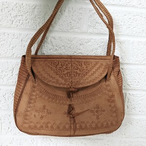 Puede incluir: Un bolso de hombro de cuero marr&oacute;n con intrincados dise&ntilde;os en relieve. El bolso tiene un cierre de solapa con una correa anudada.