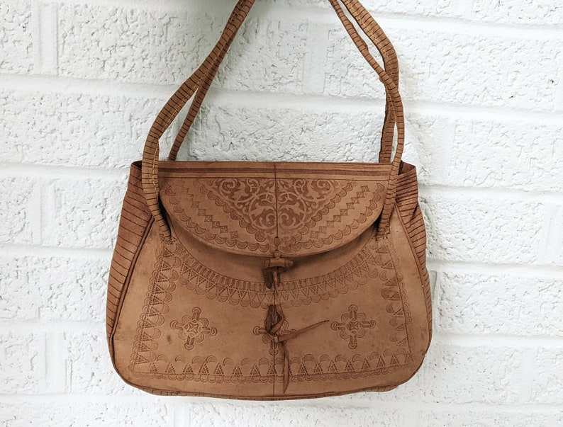Puede incluir: Un bolso de hombro de cuero marr&oacute;n con intrincados dise&ntilde;os en relieve. El bolso tiene un cierre de solapa con una correa anudada.