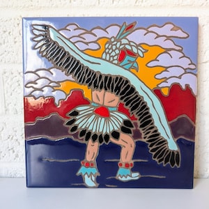 Puede incluir: Un azulejo de cerámica con una ilustración colorida de un bailarín nativo americano que lleva un tocado de plumas y un traje azul y blanco. El bailarín está rodeado de nubes y una puesta de sol.
