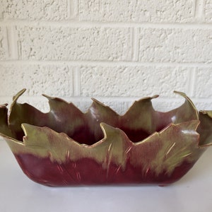 May include: A red and green ceramic bowl with a leaf design. The bowl is made of glazed pottery and has a unique, organic shape.