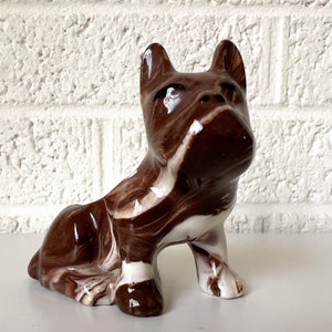 May include: A brown and white ceramic figurine of a dog sitting with its head tilted up. The dog has a marbled glaze.