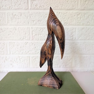 Pode incluir: Escultura de madeira artesanal de um pássaro com um pescoço longo e curvo e um bico pontudo. A escultura é feita de madeira castanha escura com padrões de grão visíveis e assenta numa pequena base angular. O pássaro está numa superfície verde.