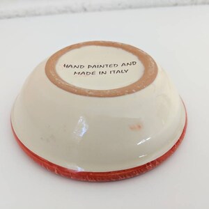 May include: A small, white ceramic bowl with a red rim. The bottom of the bowl is stamped with the text "Hand painted and made in Italy."