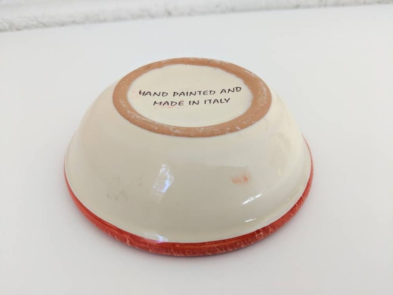 May include: A small, white ceramic bowl with a red rim. The bottom of the bowl is stamped with the text "Hand painted and made in Italy."