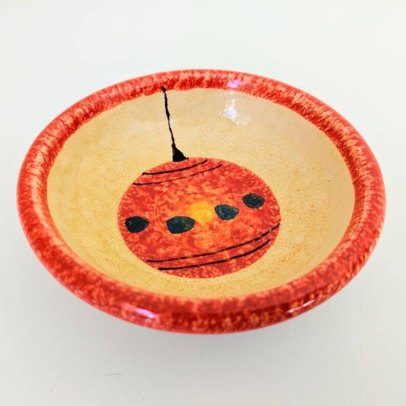 May include: A small, round ceramic bowl with a red rim and a hand-painted red and yellow ornament design. The ornament has black lines and three black dots.