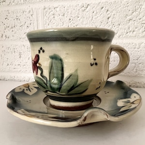 Könnte beinhalten: Ein handbemaltes Keramik-Teetassen- und Untertassen-Set mit floralem Design. Tasse und Untertasse sind beide cremefarben mit grünen Blättern, roten Blüten und schwarzen Punkten.