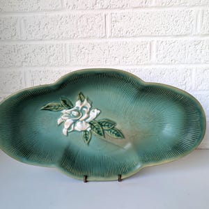 Puede incluir: Un plato de servir de cer&aacute;mica verde con un dise&ntilde;o de flor blanca en relieve. El plato tiene un borde festoneado y una superficie texturizada.