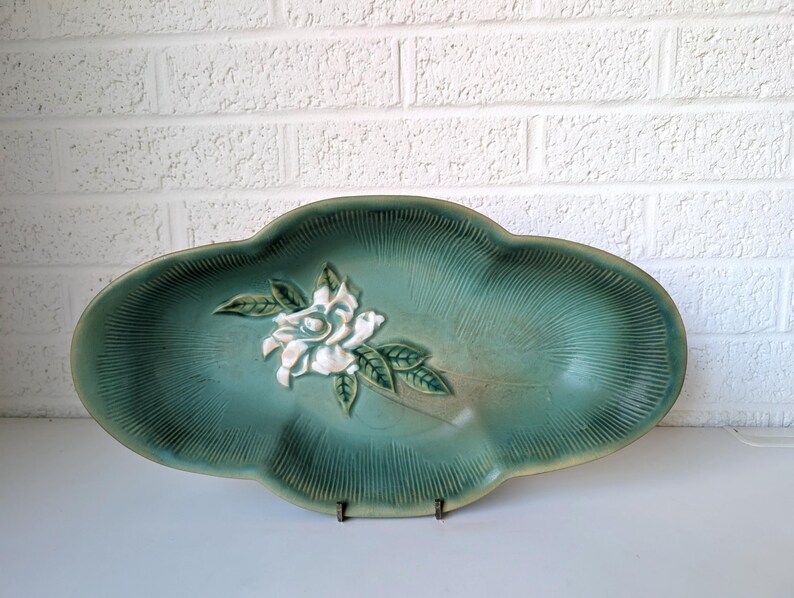 Puede incluir: Un plato de servir de cer&aacute;mica verde con un dise&ntilde;o de flor blanca en relieve. El plato tiene un borde festoneado y una superficie texturizada.