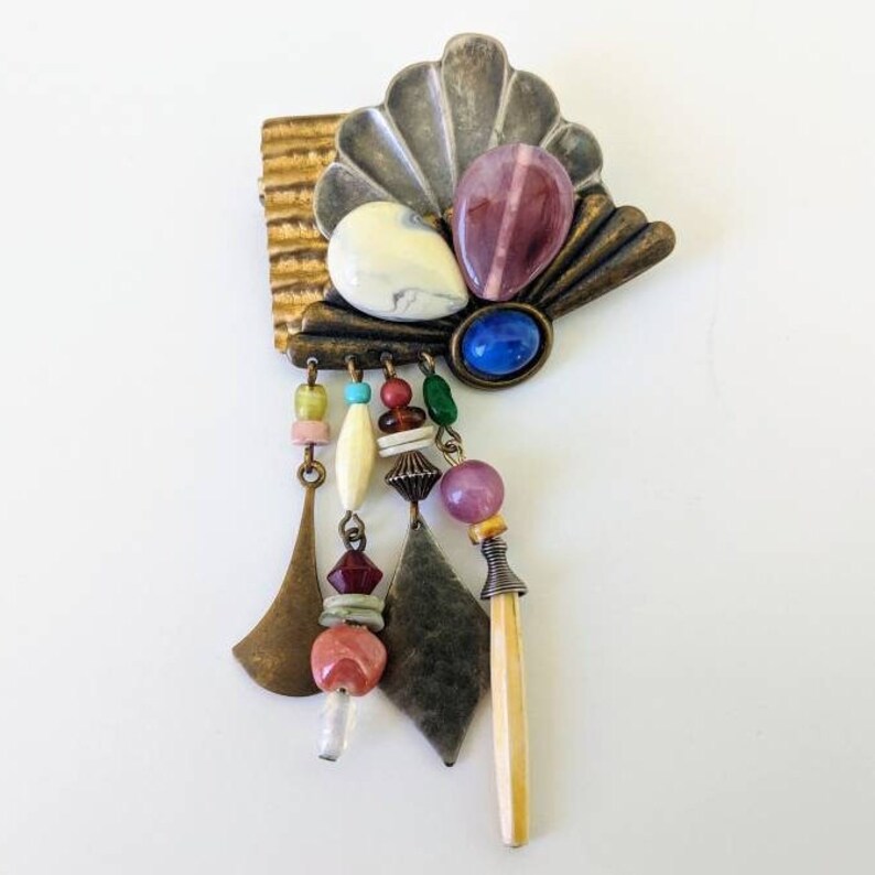 May include: A colourful, multi-textured brooch with a shell-like design, dangling beads, and various stones in shades of pink, blue, green, and white.